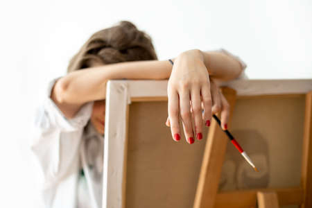 Tired artist leaned on easel, closeup. Model in white shirt with brush after hard work, resting in workplaceの写真素材