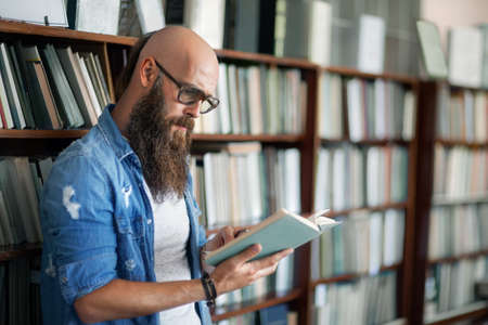 Handsome bearded man wearing glasses reading book in libraryの写真素材