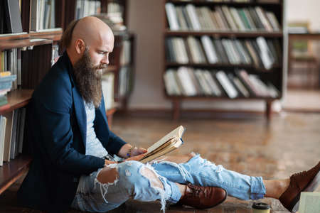 Stylish hipster man reading book sitting on floor in library or bookstore. Education, hobby conceptの写真素材