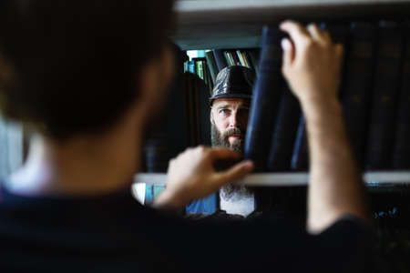 Man taking book from library bookshelf and sees bearded man. Peeping man in black hat among bookshelvesの写真素材