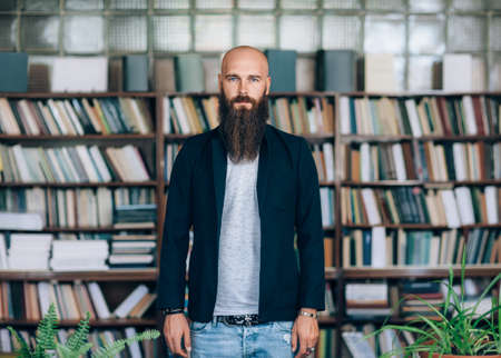 Portrait of bearded hipster man in library. Bald man with long beard stands on background of books. Education conceptの写真素材
