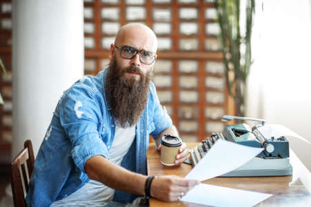 Bearded stylish writer with cup of coffe reading his novel. Modern writer in glasses working on new book in libraryの写真素材