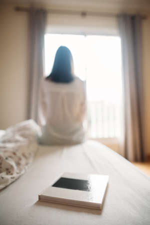 Book on bed, woman sitting in front of window in the morningの写真素材