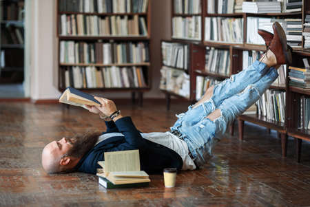 Bearded hipster man relax and reading book lies on the floor in libraryの写真素材