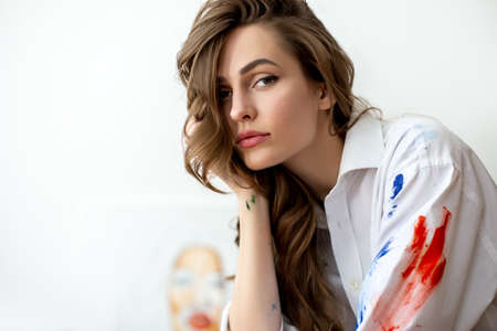 Young beautiful woman portrait on white stained with paint shirt. Thoughtful artist in studioの写真素材