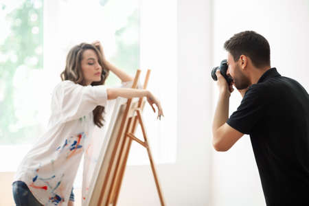 Pretty female artist posing by easel on art studio. Caucasian young woman in stained shirt on photo shootの写真素材