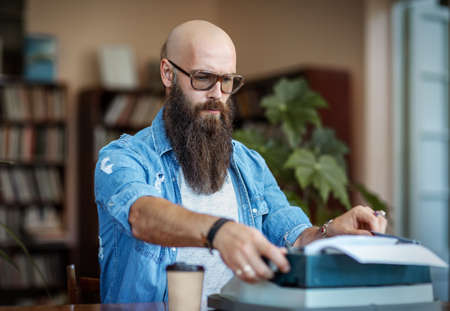 Bearded stylish writer typing on typewriter. Modern writer in glasses working on new book in libraryの写真素材