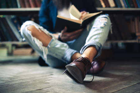 Young student man sitting on floor, reading book and relaxing in library. Focus on bootsの写真素材