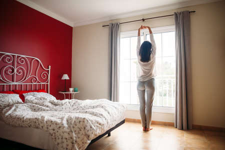 Full length portrait of happy beautiful woman stretching near window after wake up. Good morningの写真素材