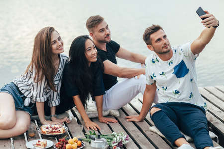 Friends making selfie sitting on pier with lake background while resting at picnic. Friendship and...