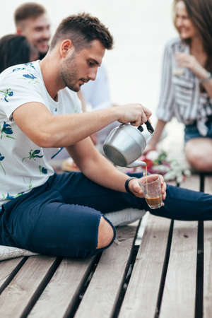 Young man pours tea into cup on picnic sitting on wooden pier. Camping food conceptの写真素材