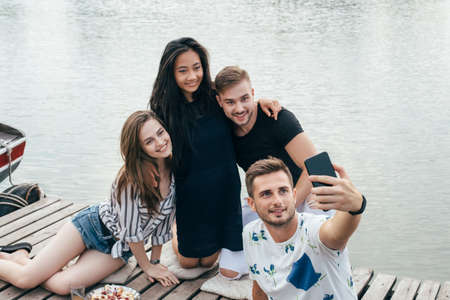 Friends making selfie sitting on pier with lake background while resting at picnic. Friendship and fun conceptの写真素材