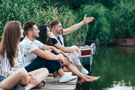 Group of friends relaxing on lake. Man in background laughs and points with his hand forwardの写真素材