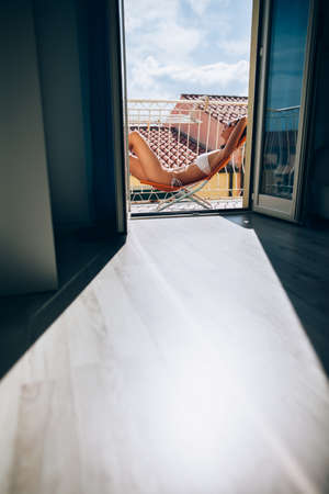 Young woman resting on sun chair and sunbathing on hotel terrace on city roofs background. Vacation and summertime conceptの写真素材