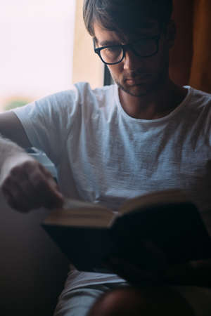 Young handsome man in eyeglasses reading book sitting near window. weekend conceptの写真素材