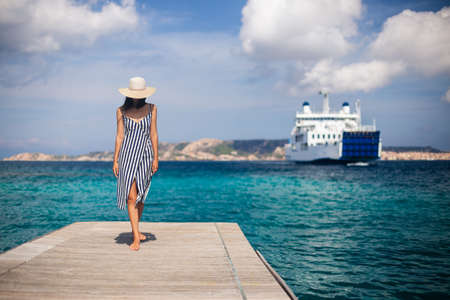 Beautiful young woman in hat walking on wooden pier on Sardinia beach, cruise ship on backgroundの写真素材