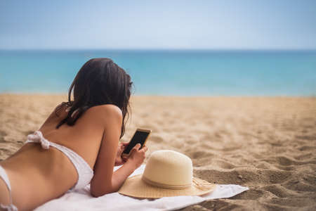 Young beautiful woman in bikini lying on tropical beach and using mobile phone. Summer vacation conceptの写真素材