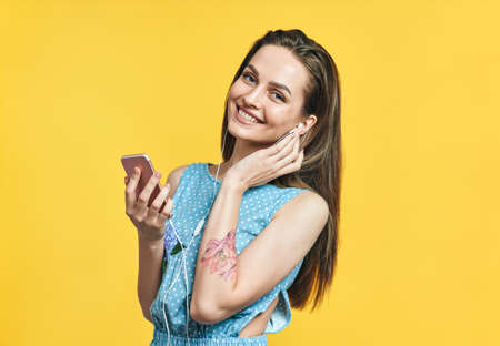 Beautiful young woman in headphones and smart phone listening music on yellow background