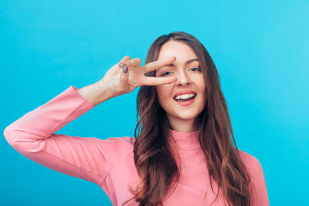 Happy beautiful woman showing peace sign near eye isolated on blue background.