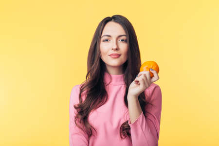 Beautiful young woman holding orange in hand isolated on yellow background. Healthy food concept