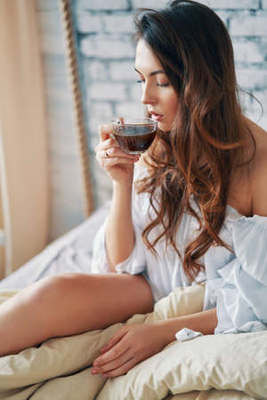 Young beautiful woman drinking hot coffee sitting in bed and enjoying her good morning. Relax and carefrre conceptの写真素材