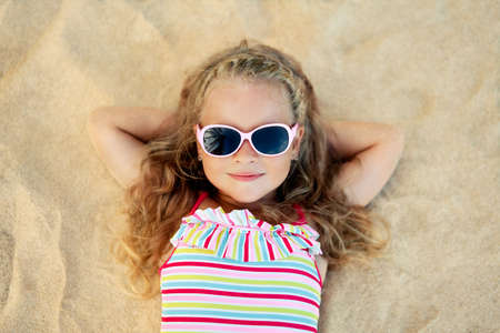 Top view of pretty little blonde girl in sunglasses lying on sandy beach during summer vacationの写真素材