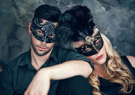Beautiful couple in mysterious black mask sitting on floor in studioの写真素材