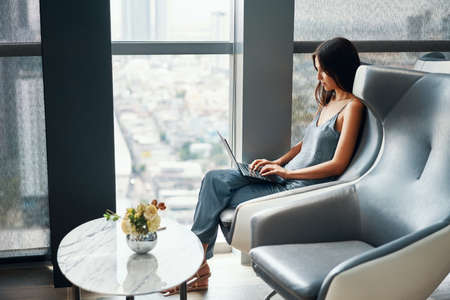 Young woman working on laptop while sitting in a big comfortable chair at modern interior