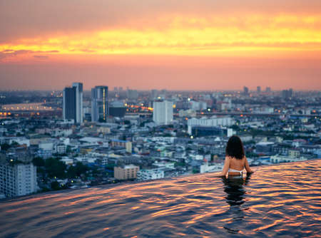 Young woman relax in swimming pool on roof top during amazing sunset and enjoy cityscapeの写真素材