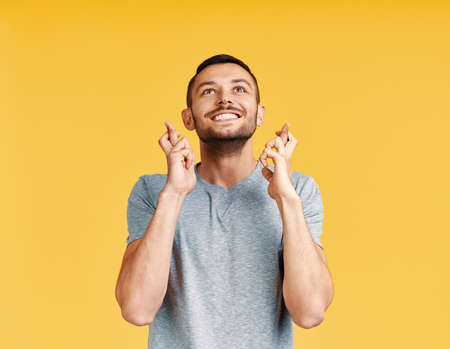 Young man making a wish and ask for good luck with crossed fingers isolated on yellow background