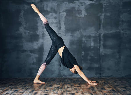 Young sporty woman practicing yoga, doing one legged downward facing dog pose on dark background