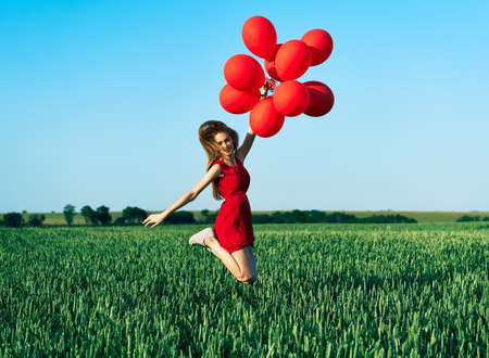 Young happy woman jumping with red balloons on green summer field