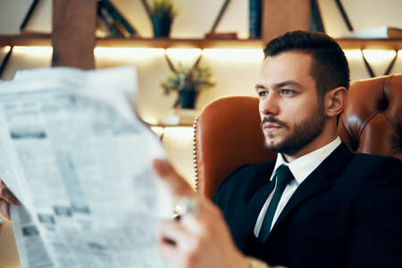 Confident young businessman reading newspaper and latest news while sitting in armchairの写真素材