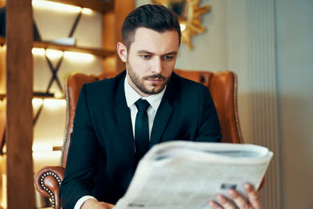 Confident young businessman reading newspaper and latest news while sitting in armchairの写真素材