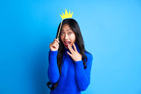 Surprised beautiful woman hold a paper craft crown on a stick on blue background. Party, emotions conceptの写真素材