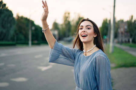 Young pretty woman waving raised hand hitchhiking or welcome friends on city background. Hello, greeting conceptの写真素材