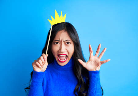 Angry screaming woman with paper crown on stick isolated on blue background. Party, emotions concept                の写真素材