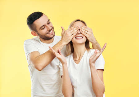 Guess who? Surprise for her. Young man covering eyes of his girlfriend isolated on yellow background