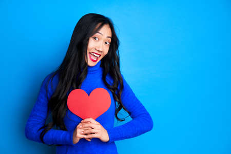 Smiling happy woman holding big paper heart in her hands isolated on blue background. Love conceptの写真素材