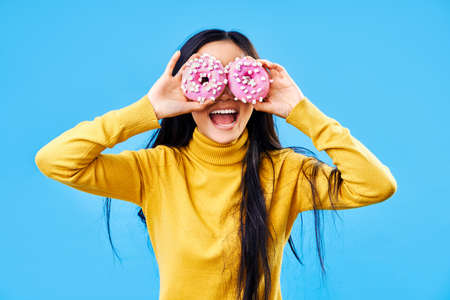 Attractive woman having fun with donuts. Girl showing doughnuts in front of her eyes on blue background. Tasty food, diet conceptの写真素材
