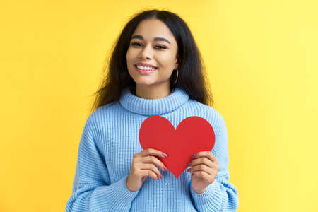 Portrait of pretty smiling black woman holding big paper heart in her hands on yellow background