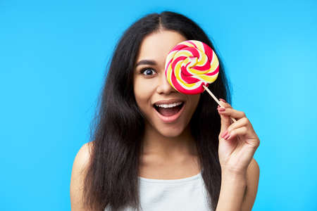 Excited happy black woman covers her eye with colorful lollipop isolated on blue backgroundの写真素材