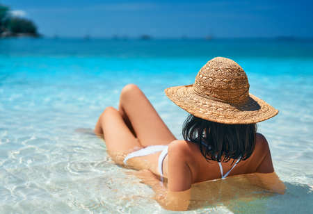 Back view of young woman in straw hat and white bikini relax in turquoise sea on tropical paradise...