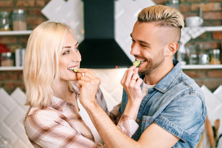 Beautiful young couple is feeding each other and smiling while cooking in kitchen at homeの写真素材