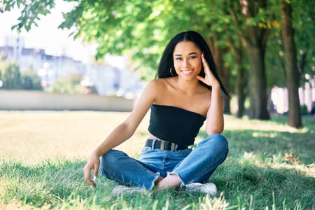 Pretty smiling afro american female posing in park sitting on green grass.