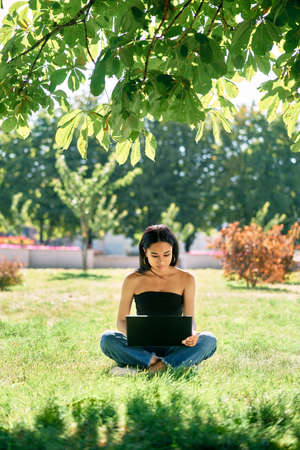 Millennial afro girl working on laptop sitting on grass in park