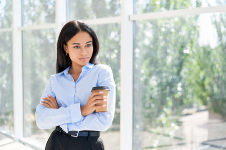 Pretty black business woman holding cup of coffee in hand posing on panoramic windows background in...