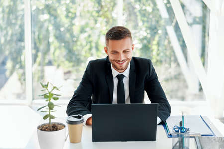 Handsome smiling businessman in elegant suit working on laptop in a bright modern officeの写真素材