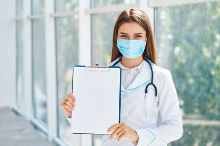 Confident female doctor wearing medical mask showing clipboard with diagnosis in hospital backgroundの写真素材