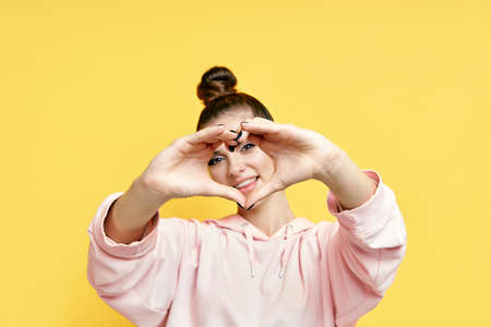 Young smiling pretty woman making heart symbol by hands on yellow background